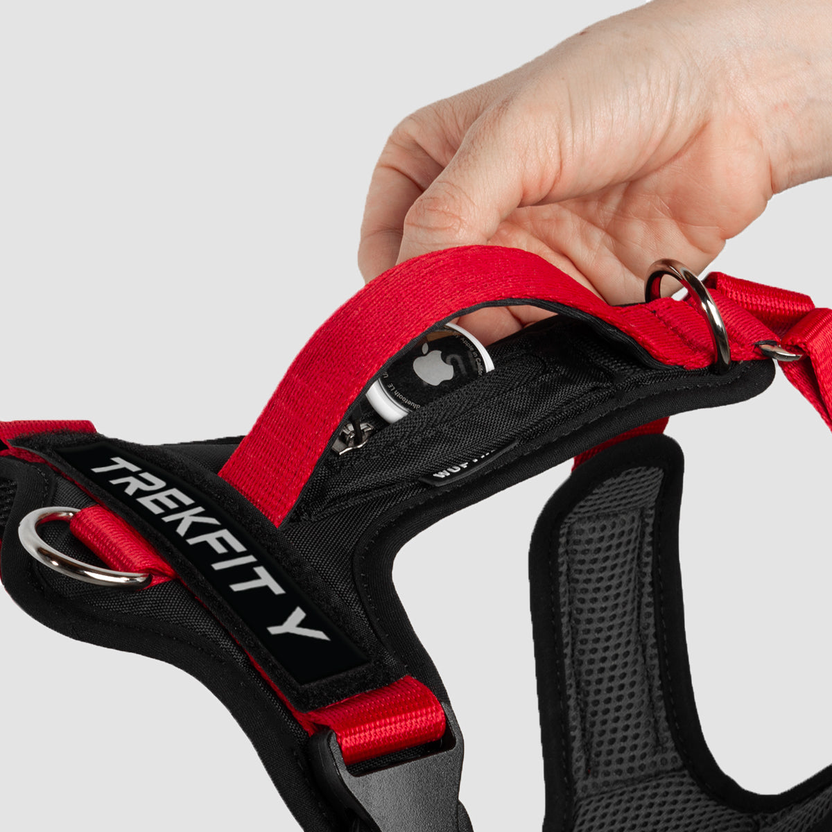 Un Arnes en forma de Y en color rojo con negro, visto desde arriba, se puede observar la manija de control, las argollas de atras, una mano sosteniendo un localizador AirTag dentro de un bolsillo discreto, y el espacio con parche de velcro