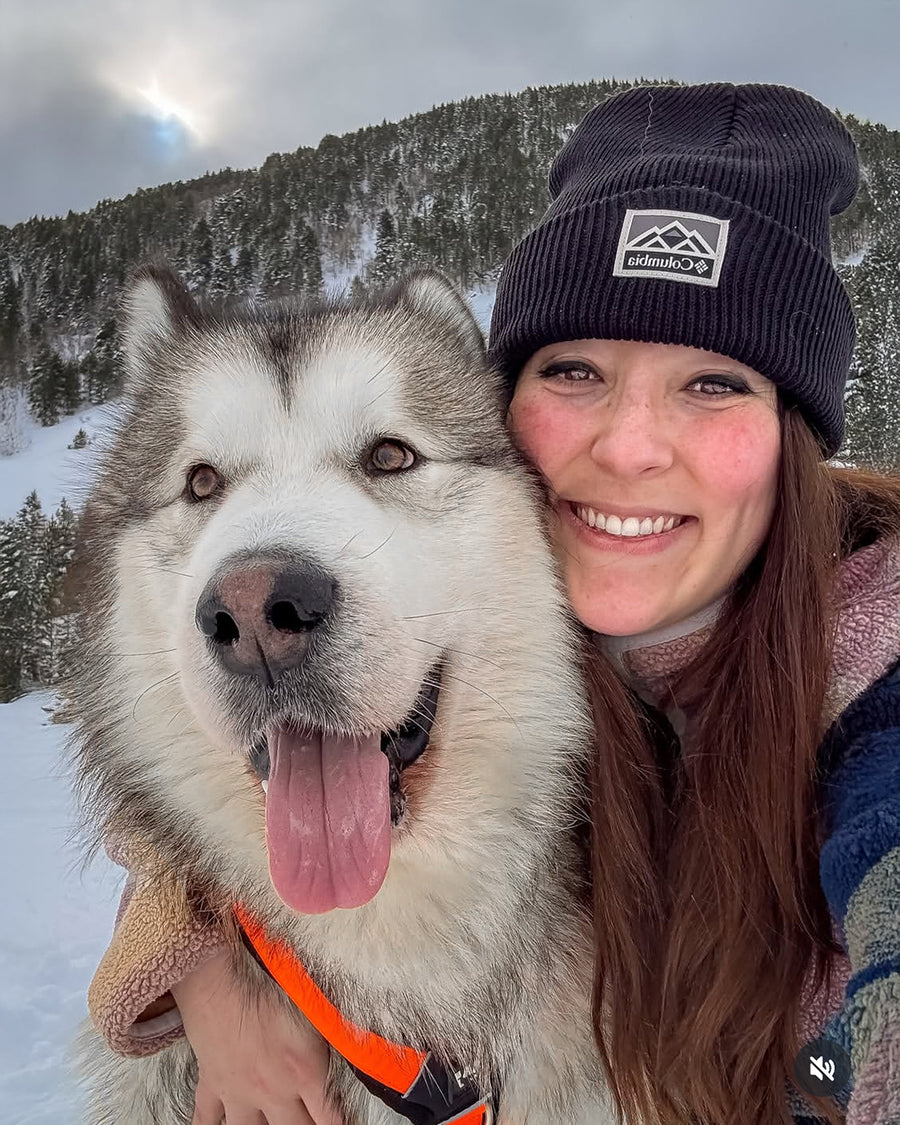Un Alaskan Malamute con la lengua afuera junto a una mujer mirando de cerca al a camara, con un arnés naranja. Atras una montaña nevada con pinos.